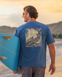 Man holding a surfboard at the beach & wearing a blue shirt with a tribal shark design on the back