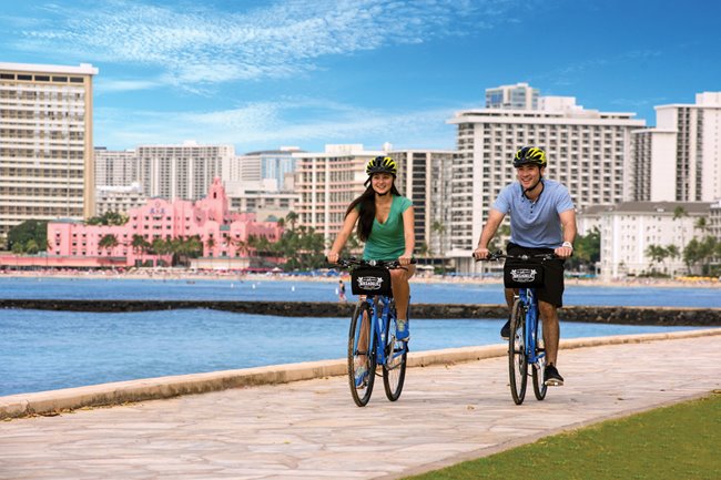 Couple biking along Waikiki with the ocean and the pink Royal Hawaiian Hotel in the background.