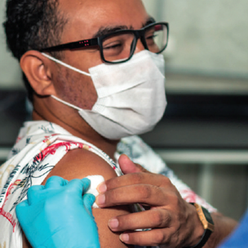 Person wearing glasses & face mask with a cotton pad being applied to his shoulder area by a healthcare professional wearing blue gloves.