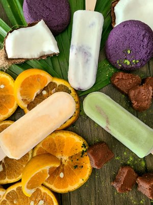 Orange, pastel blue, and white popsicles laying flat next to orange slices, ube cookies, and pieces of coconut.