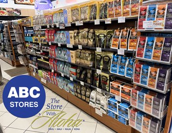 An aisle in an ABC store filled with various flavors and brands of coffee on shelves. On the floor, there's the ABC Stores logo and text "The Store With Aloha - OAHU . MAUI . KAUAI . KONA".