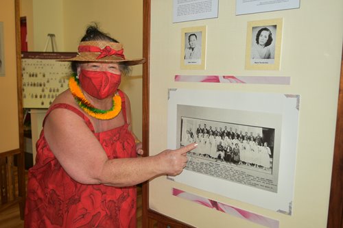 A woman in a red muumuu, mask, hat & lei pointing at a black & white photo on one of the Hawaiian culture displays.