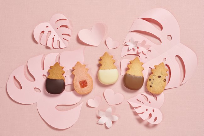5 Honolulu Cookie Company pineapple-shaped shortbreads on pale pink cutouts of tropical leaves.
