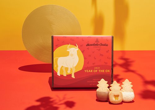 3 pineapple-shaped cookies from Honolulu Cookie Company upright in front of the 2021 red & yellow Year of the Ox packaging.