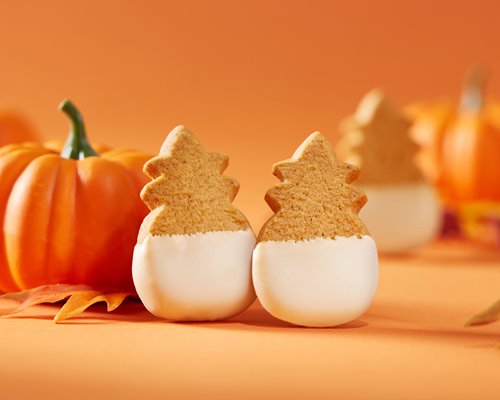 Honolulu Cookie Company's pineapple-shaped shortbread cookies dipped in white chocolate next to a mini pumpkin against an orange backdrop.