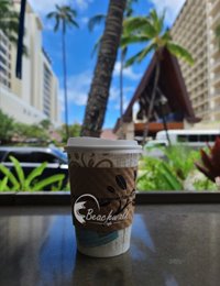 Takeaway coffee cup with the Beachwalk Cafe logo sitting on a table with palm trees and a building in the background.