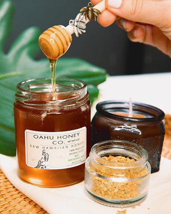 Oahu Honey Co. labeled jar next to a candle with a hand holding a honey dipper that is lifting some of the honey out of the jar. lifting a