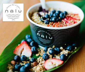 Acai bowl in a paper cup that reads Nalu Health Bar & Cafe. The cup is placed on a long green leaf which has strawberry slices, blueberries and oats on top of it.