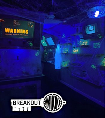 A dark escape room with blue lights and glow-in-the-dark highlights. The screen reads "WARNING." The picture is overlaid with a key-like logo that reads "Breakout Waikiki."