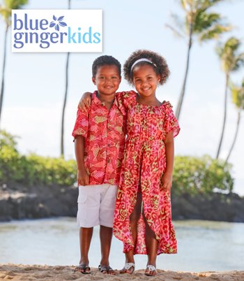 Two children wearing bright tropical clothes stand on the beach, surrounded by sand and the sparkling ocean waves.