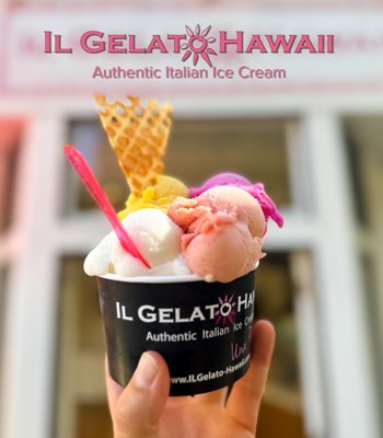 Someone holding a paper cup with white, pink and yellow scoops of ice cream. There is a neon pink spoon in the ice cream and a triangular waffle cone piece. Text on the picture reads IL Gelato Hawaii, Authentic Italian Ice Cream